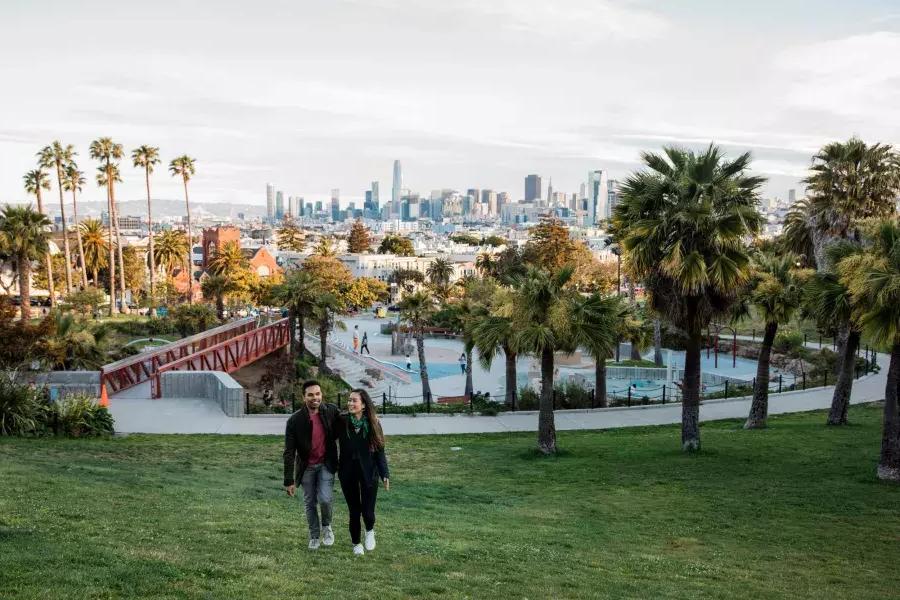 Um casal caminha em direção à câmera 和 Dolores Park e o horizonte de 贝博体彩app atrás deles.