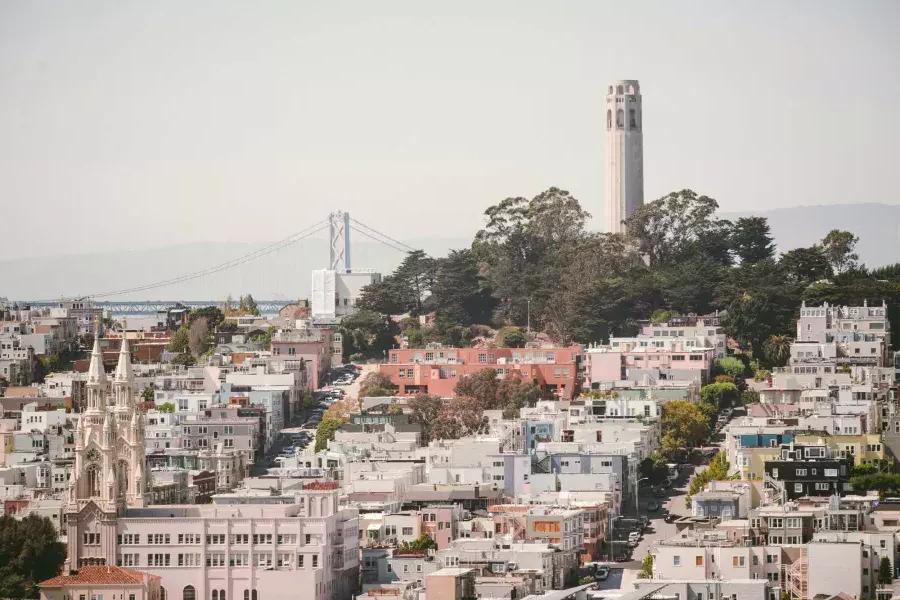 샌프란시스코의 比特塔(Coit Tower)는 배경에 베이 브리지(Bay Bridge)가 있고 전경에 집들로 뒤덮인 언덕이 있는 사진입니다.