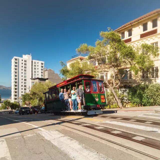 Powell and Hyde cable car going up the street with smiling people inside on a sunny day.